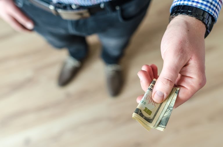 A close-up of a man's hand holding rolled US dollar bills, offering cash.